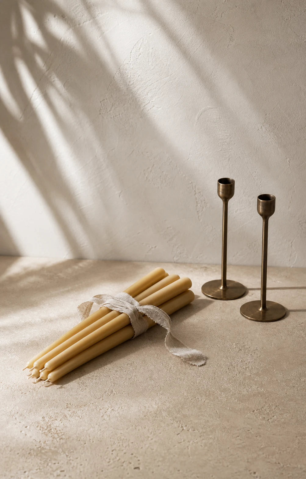 Six pale honey beeswax tapers tied with linen ribbon beside two brass holders